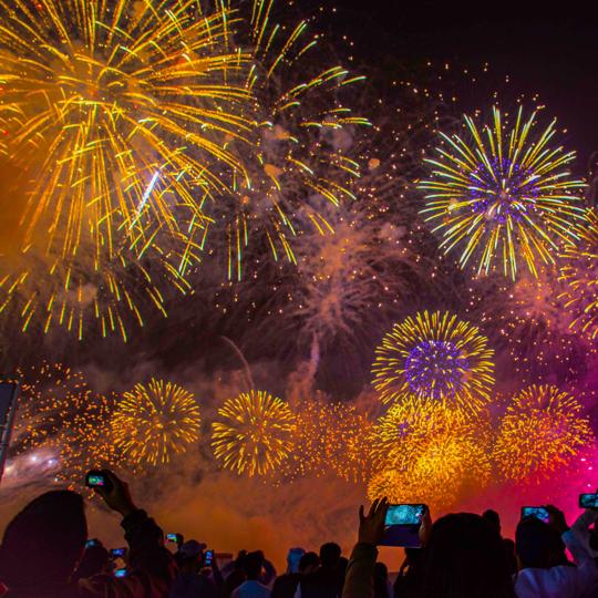 Imagem ilustrativa, noite com fogos de artificio coloridos, e pessoas fotografando o céu.